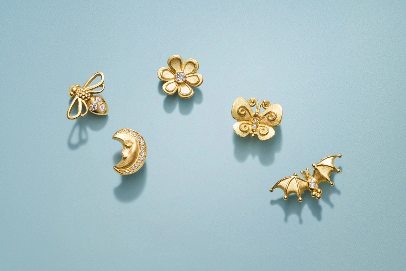 Five gold and diamond-studded charms shaped like a bee, flower, moon, butterfly, and bat on a blue background.