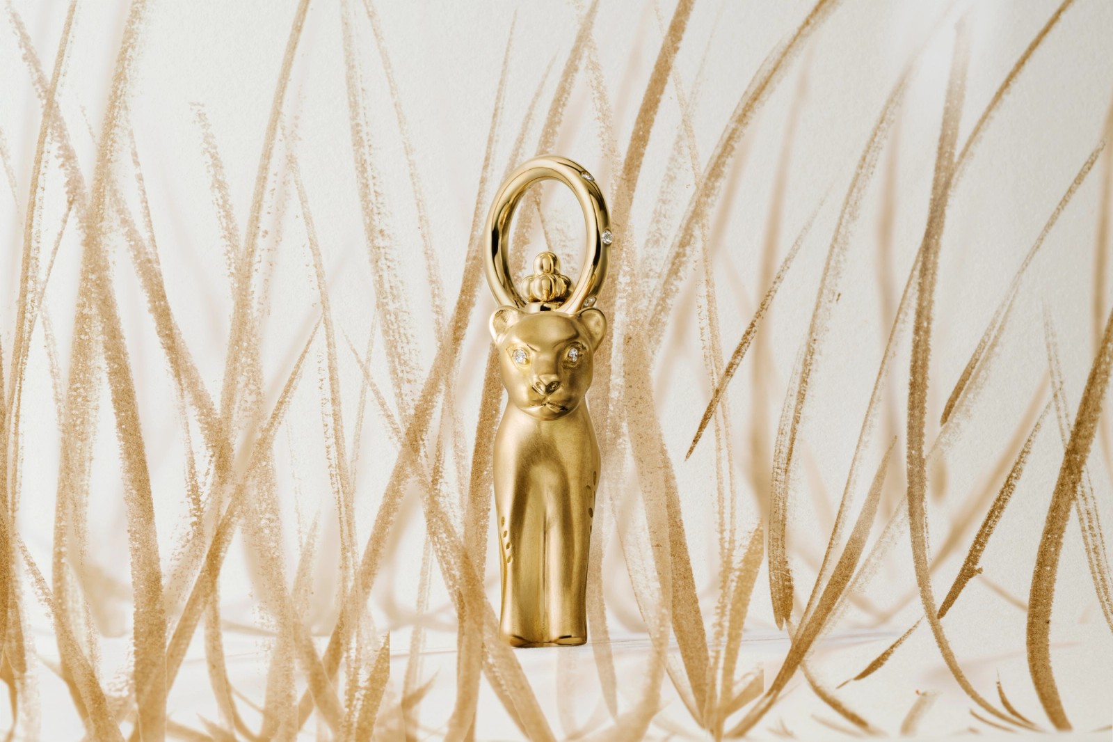 Golden panther-head object with gem eyes and a ring loop, against a light background with brown brushstrokes.