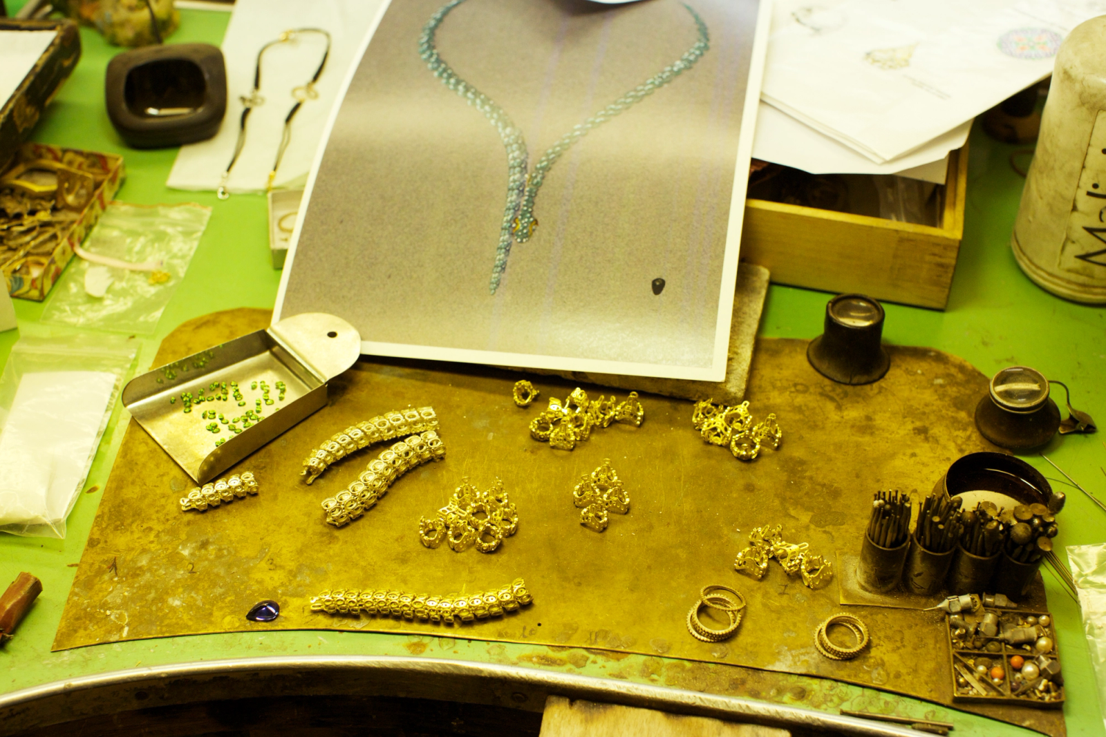 Jeweler's workbench with gold components, green beads, tools, and a necklace design sketch.