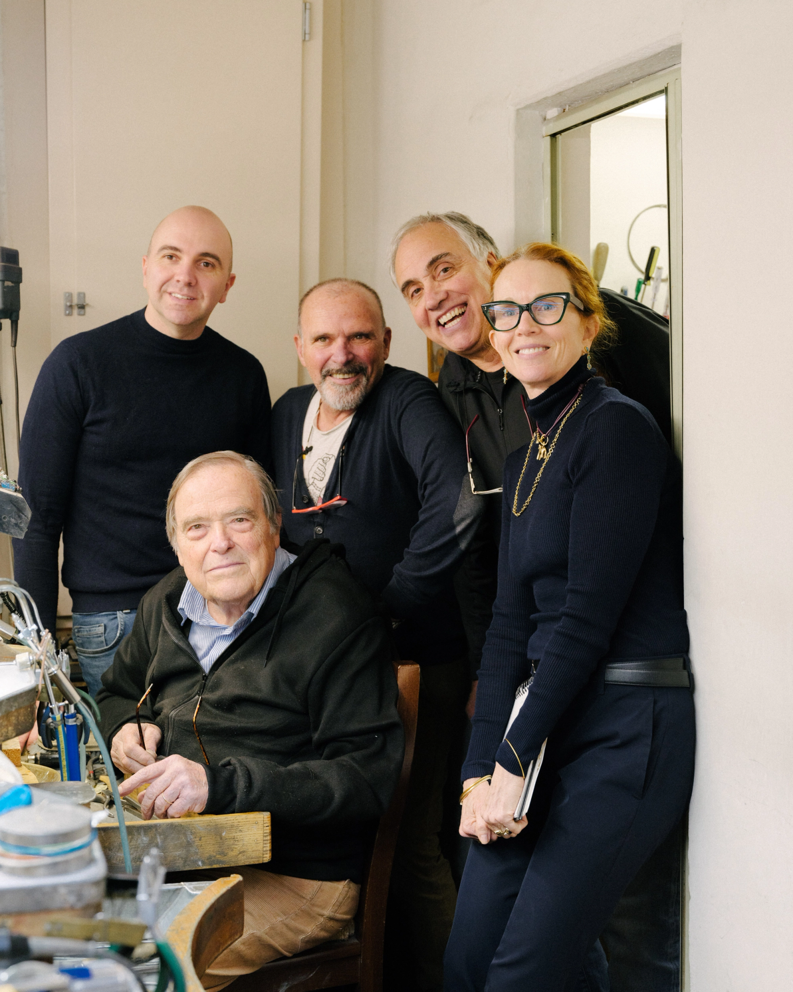 Five smiling people in a workshop, an older man seated at a workbench with four others standing behind him.