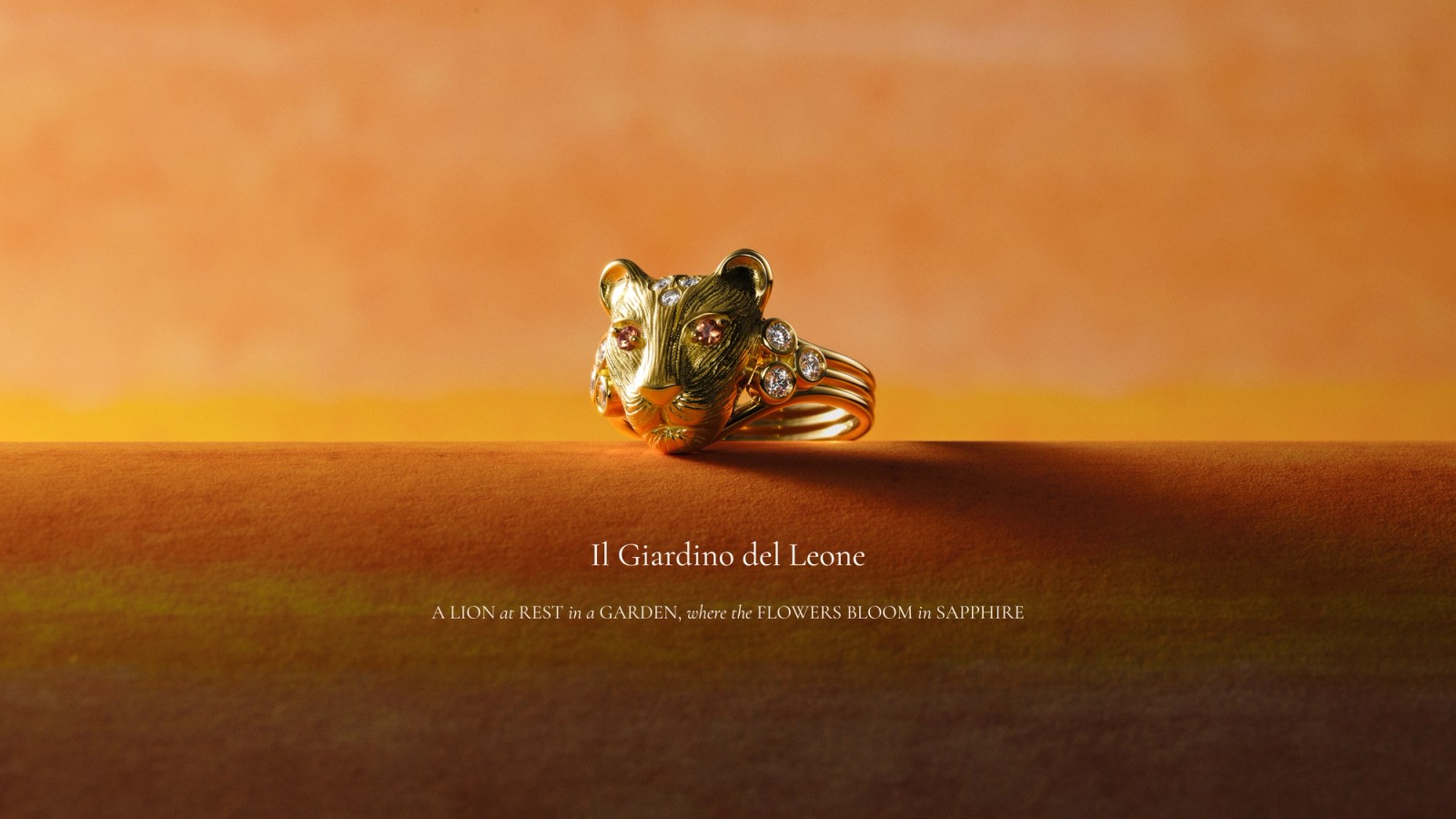 Gold lion head ring with pink and clear gemstones on an orange-brown background. Text: "Il Giardino del Leone" and "A LION at REST is a GARDEN, where the FLOWERS BLOOM is SAPPHIRE."