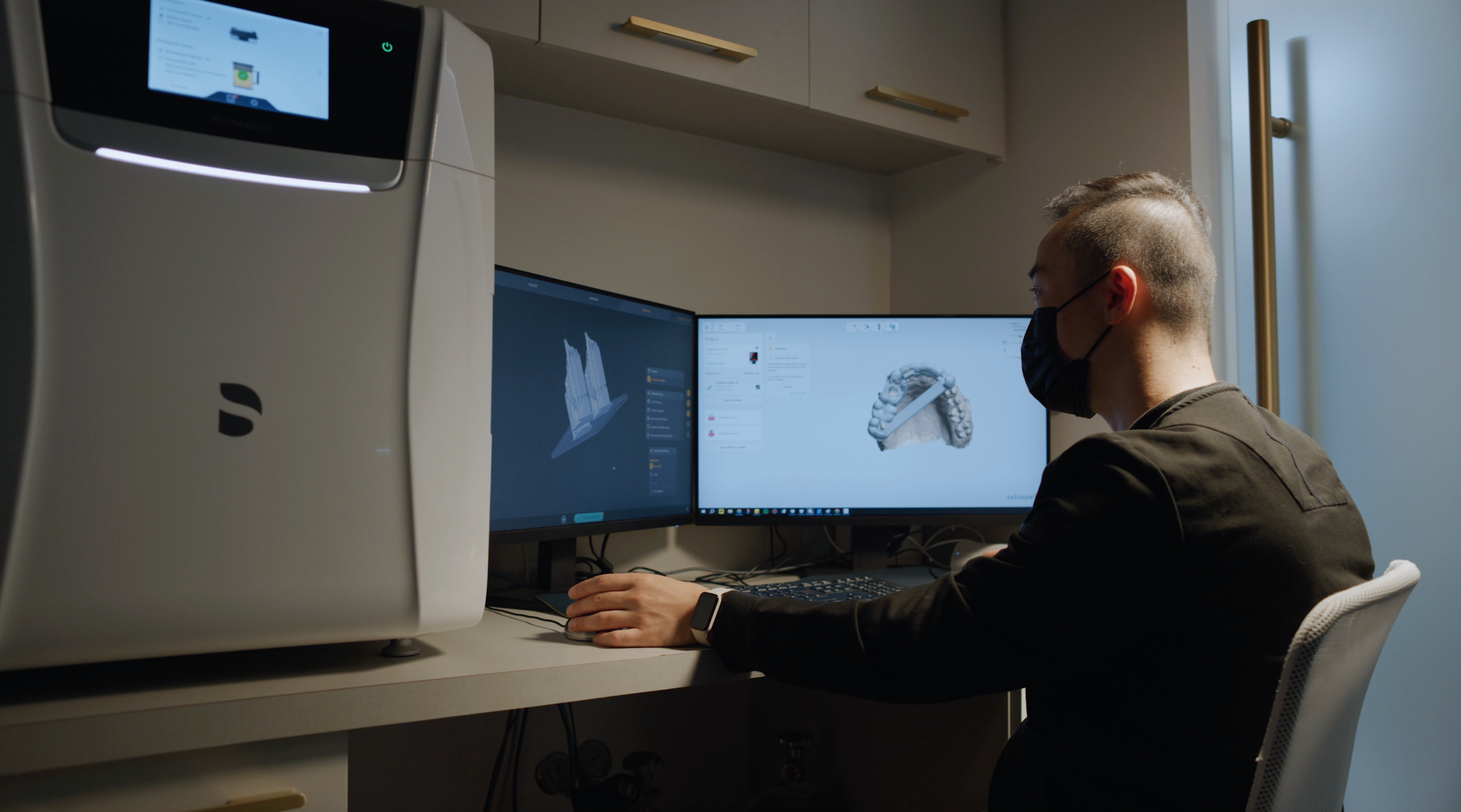 Photograph of a dental 3D printer sitting on a work bench next to dual monitors displaying 3D renders of teeth, an operator sits at the desk with their hands on the keyboard and mouse.