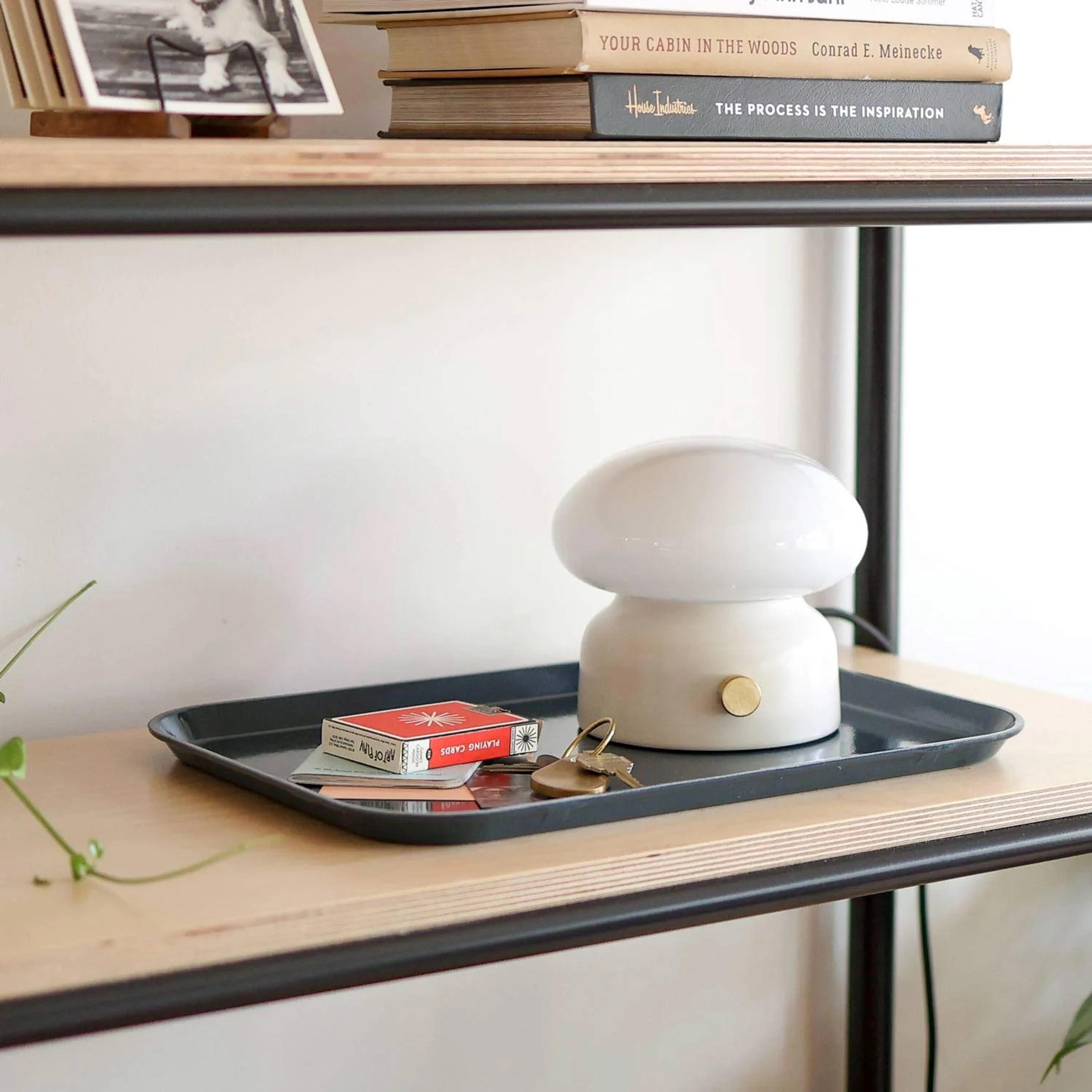 A white mushroom-shaped lamp on a black tray with keys and a deck of cards, placed on a wooden shelf. Above, books and a photo are displayed on another shelf.