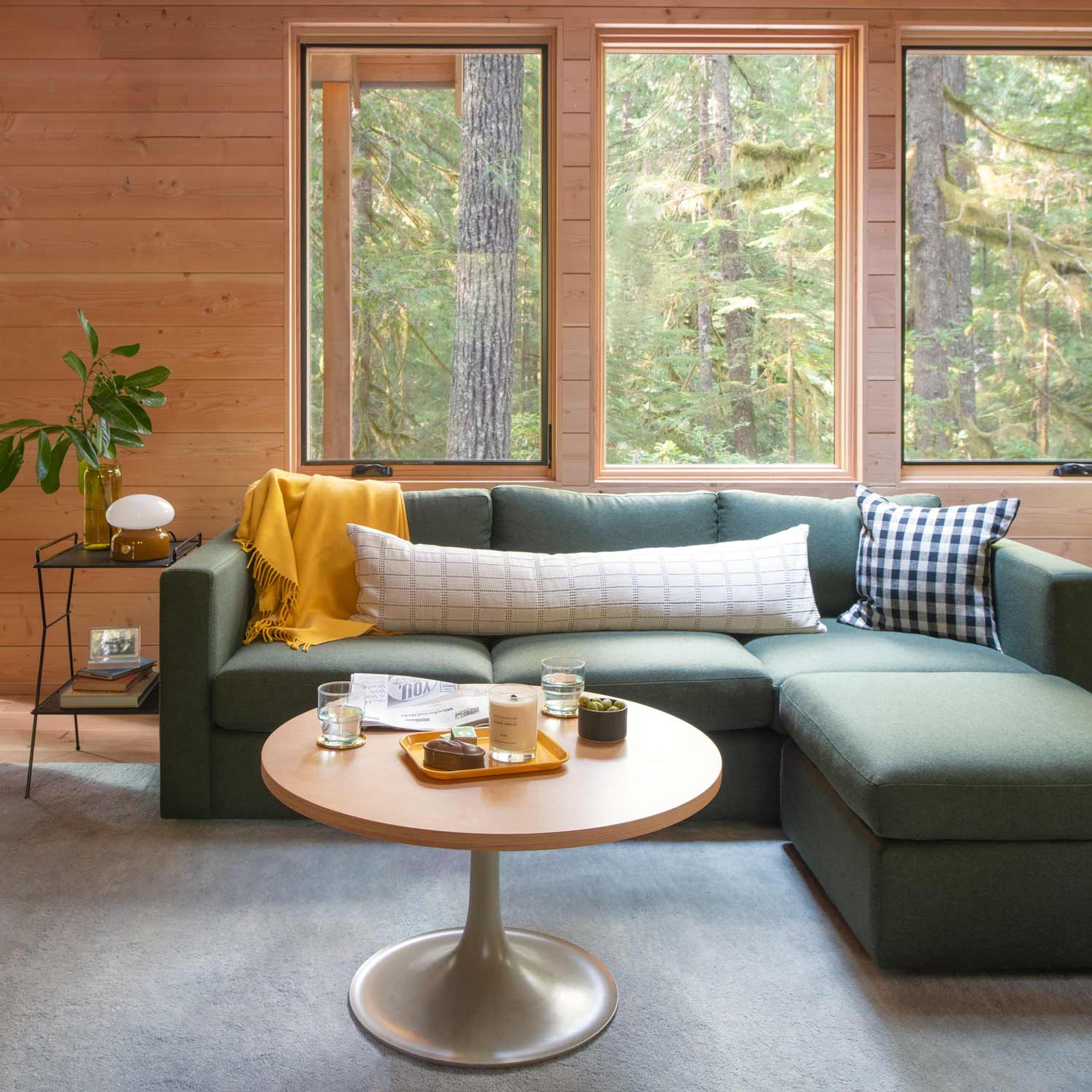 A cozy living room with a green sectional sofa adorned with a yellow throw and patterned pillows, set against large windows overlooking a forest. A round wooden coffee table holds candles and glasses, and a side table features a lamp and plant.