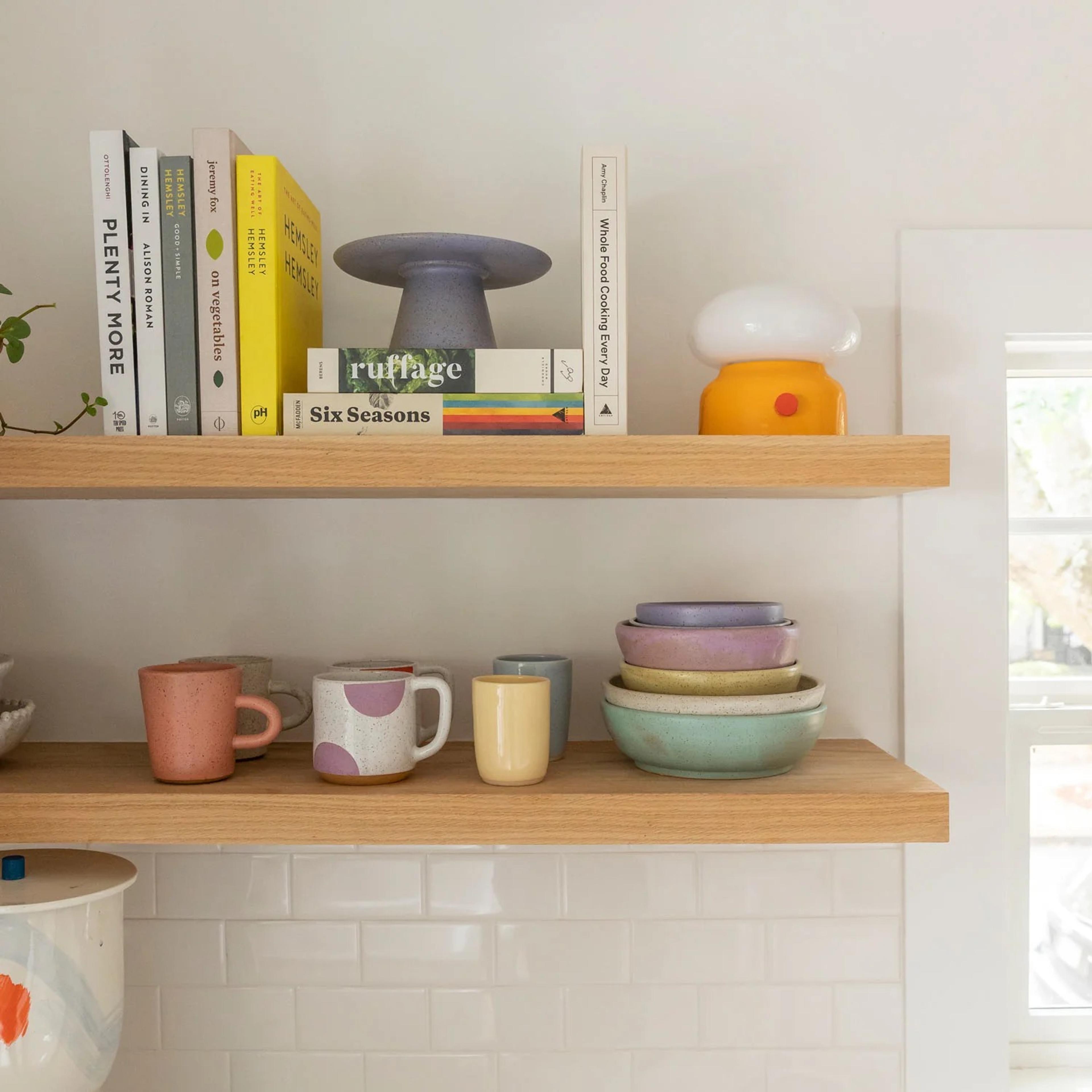 Wooden shelves with colorful books, a purple pedestal, and an orange mushroom-shaped lamp on the top shelf. The lower shelf holds various mugs and stacked pastel bowls, set against a white tiled wall.