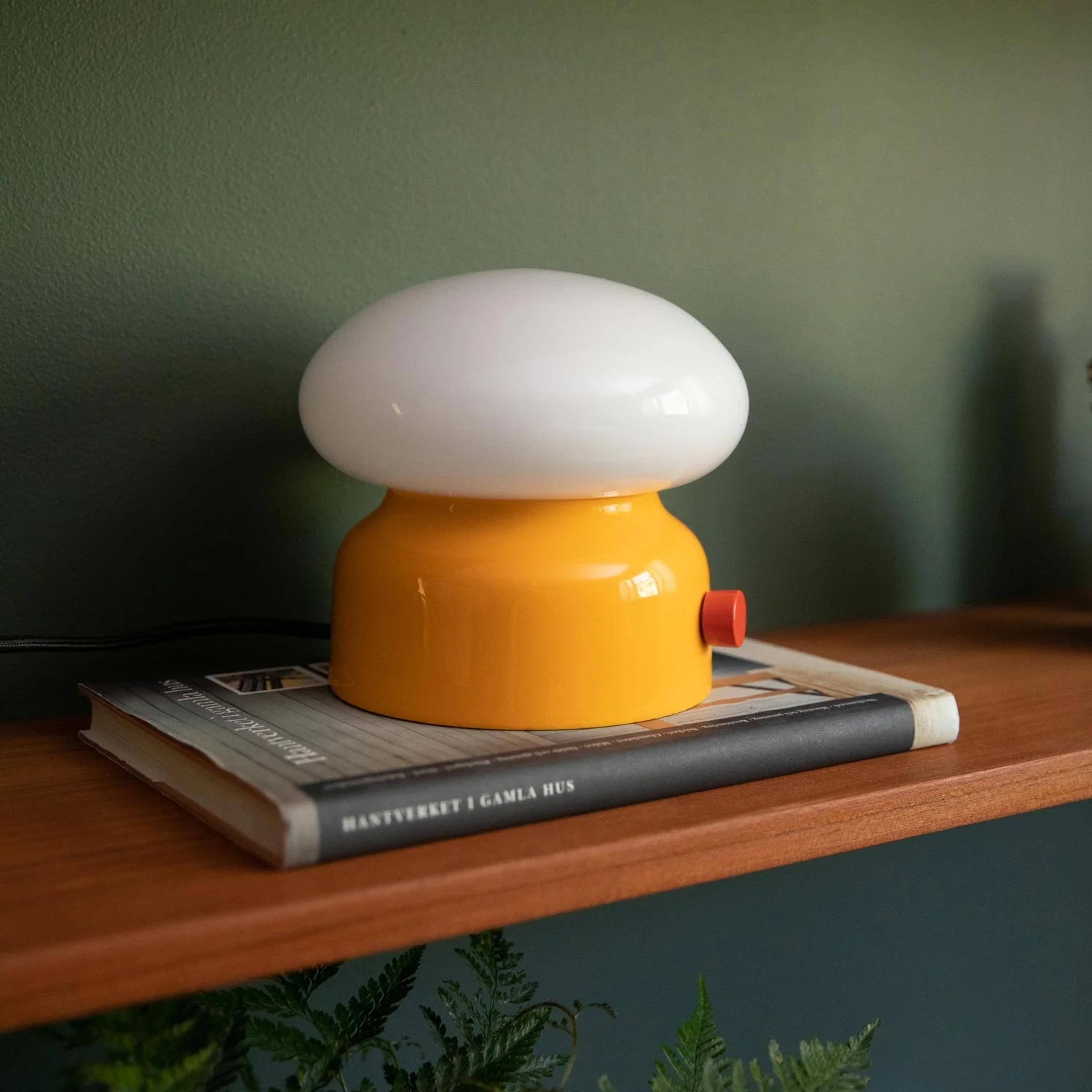 A yellow mushroom-shaped lamp with a white top sits on a book on a wooden shelf against a green wall.
