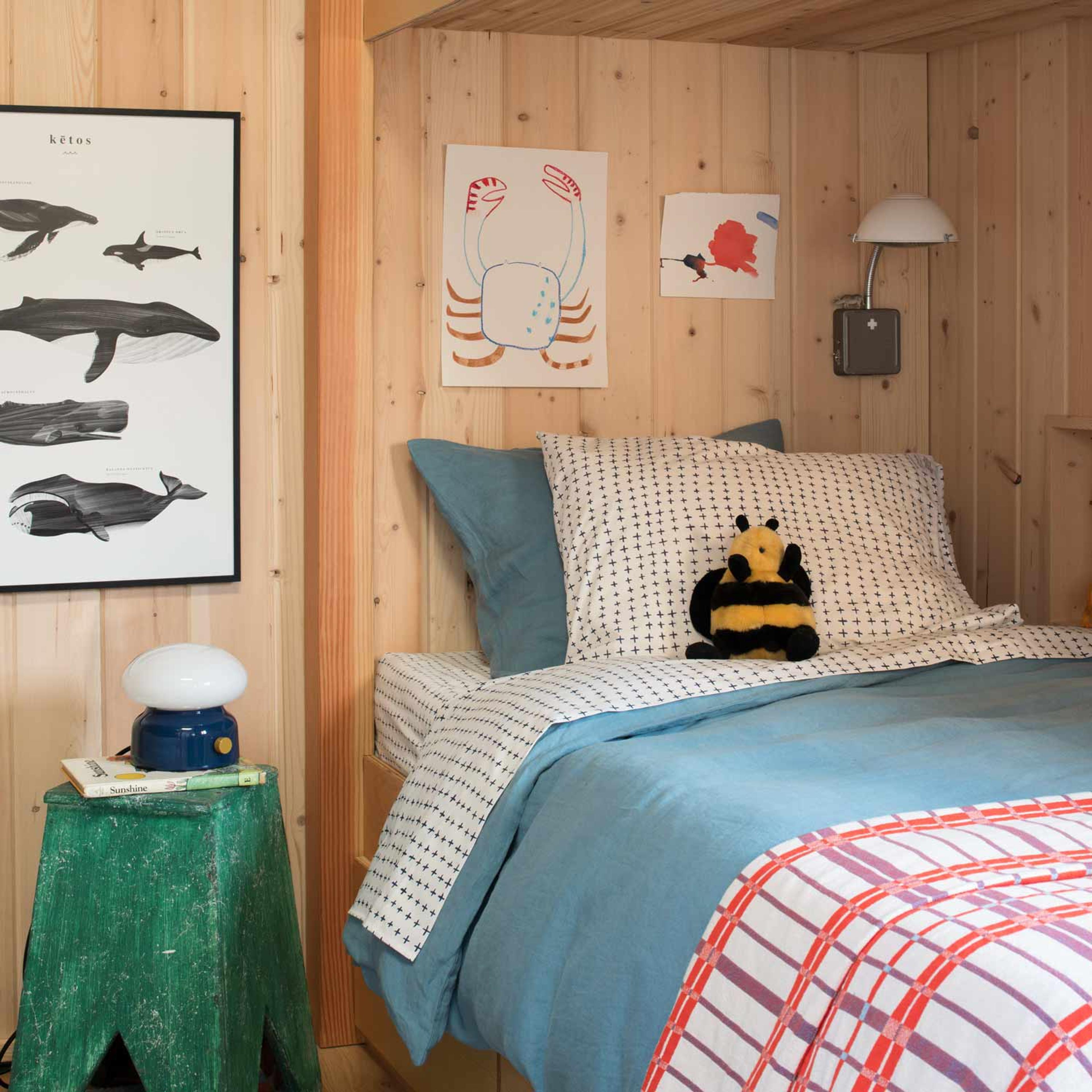 Cozy bedroom with wooden walls, a bed with blue and white bedding, a plush bee toy, and wall art of marine life and a crab. A green stool holds books and a lamp.