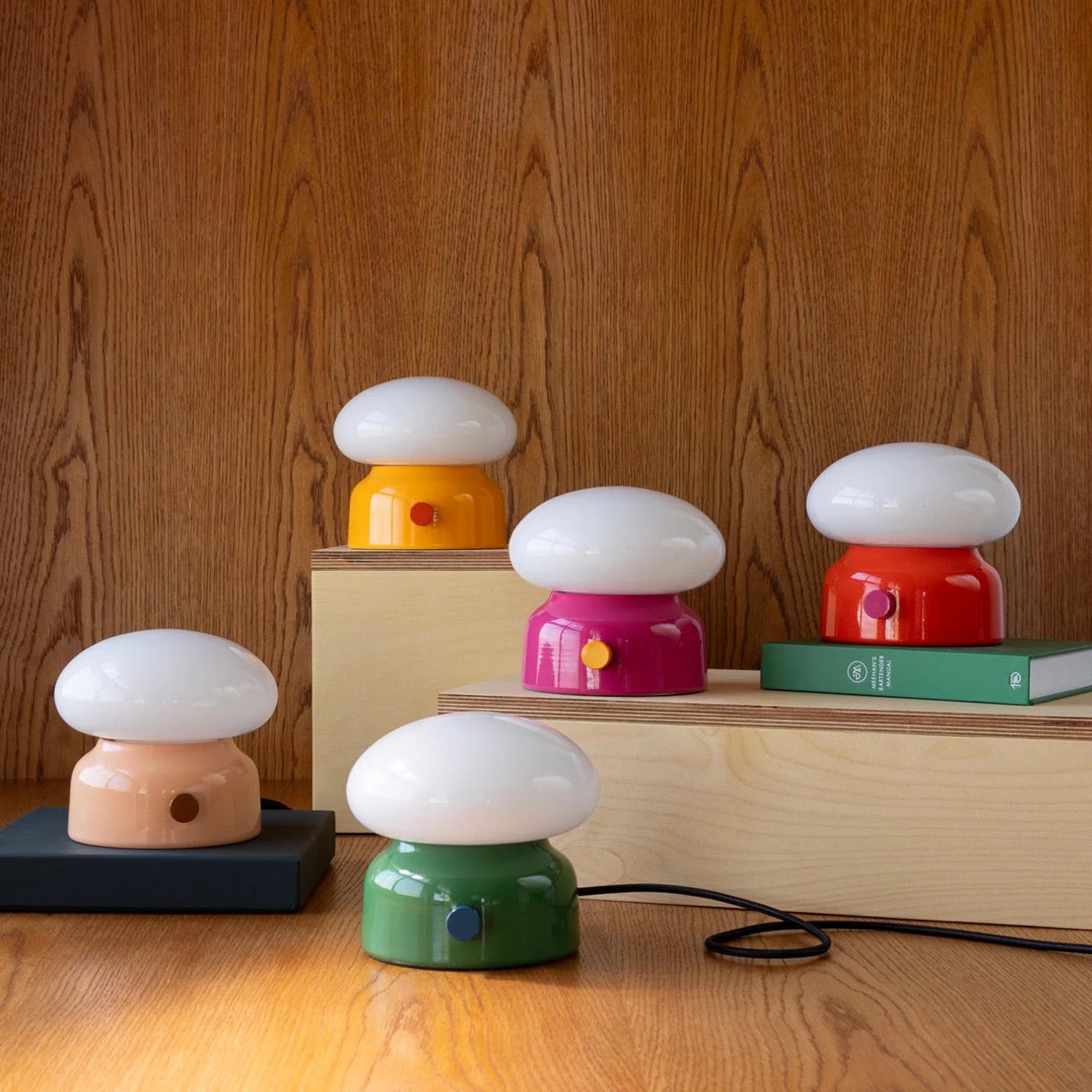 Colorful mushroom-shaped lamps in pink, orange, green, and red, placed on wooden surfaces with books, against a wood-paneled background.