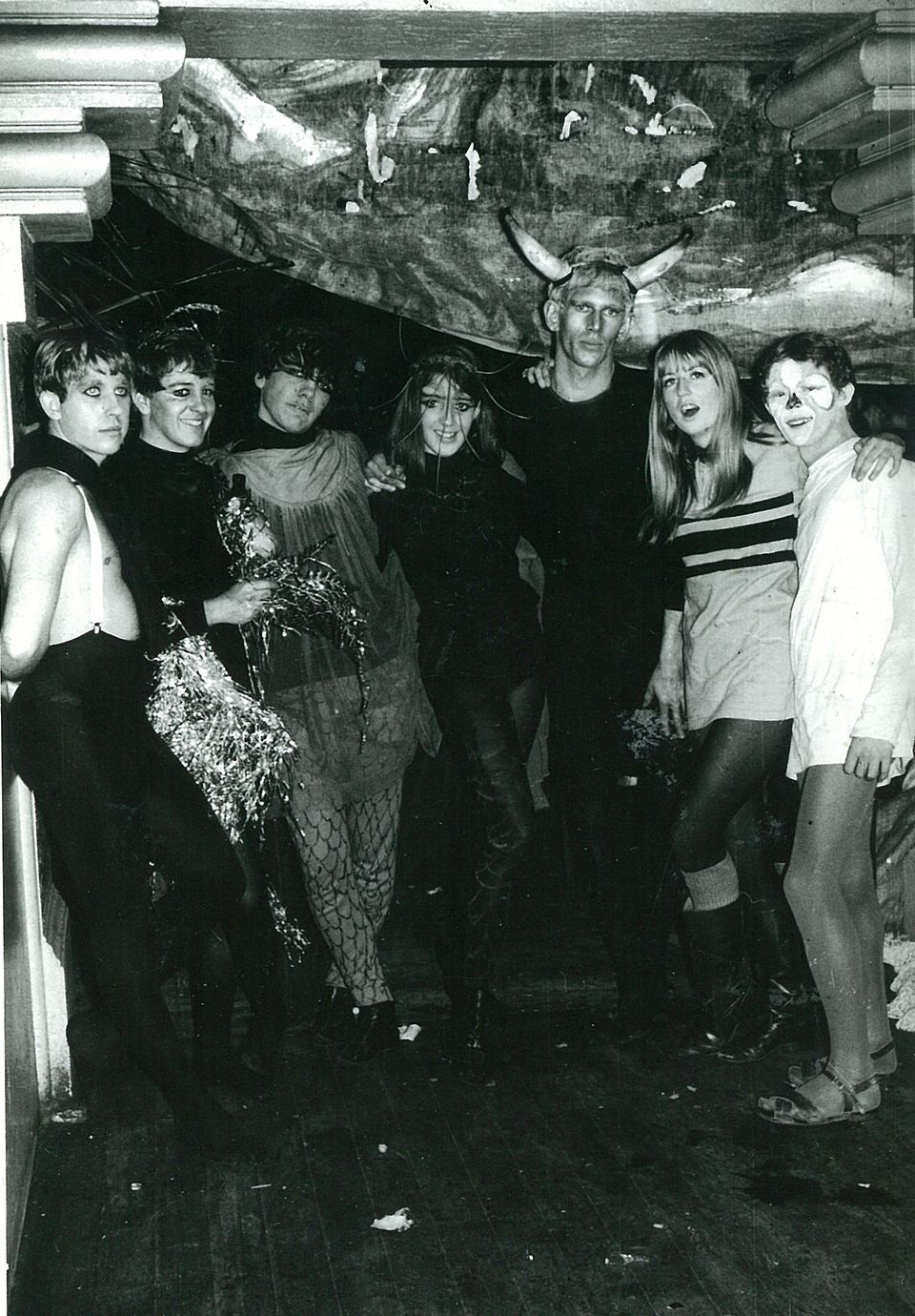 A black and white image of 7 young adults in dress up costumes standing in a semi-circle with their arms around each others shoulders 