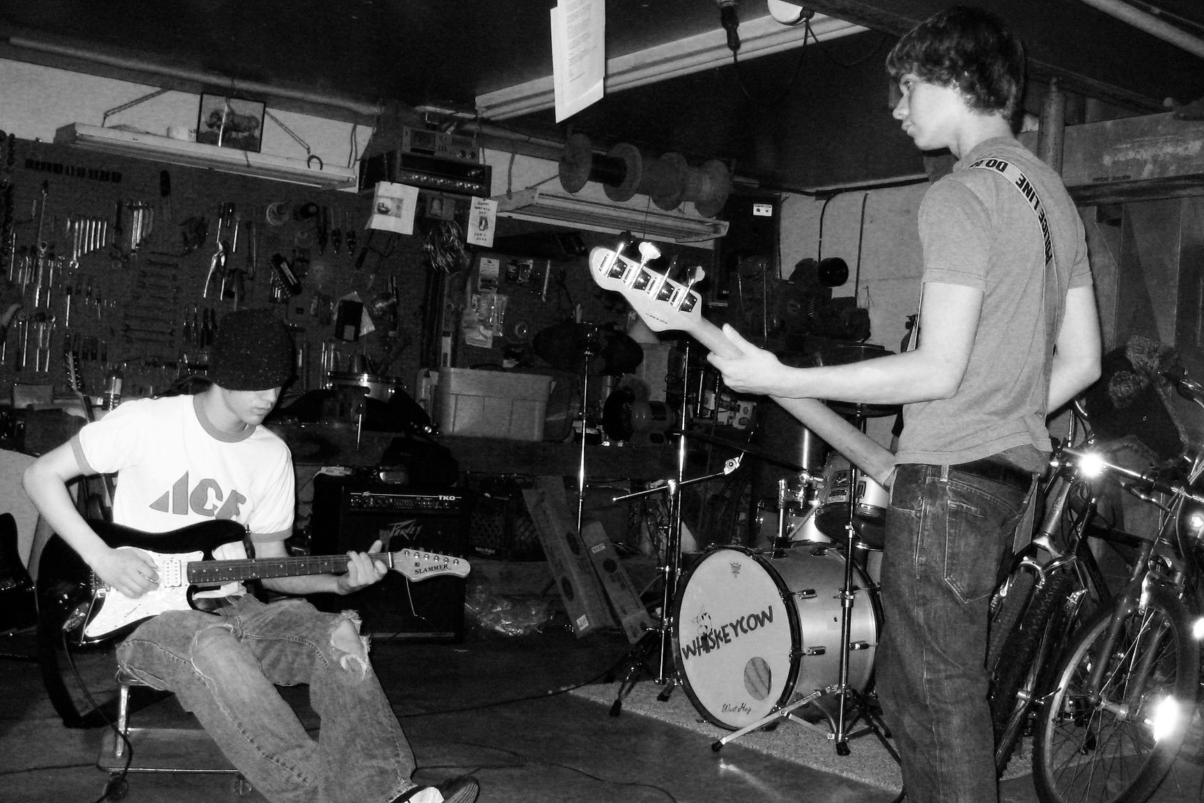 A black and white image of two male teenagers dressed in baggy jeans and t-shirts, playing guitar in a residential garage with a drumkit it the background