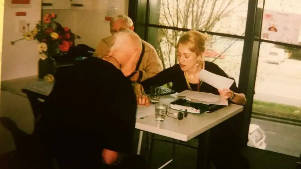 Yellowed photograph of a blonde woman seated at a desk in an office holding papers and leaning over the table to point at a piece of paper in front of a man with white hair seated opposite who is reading whats written on the piece of paper