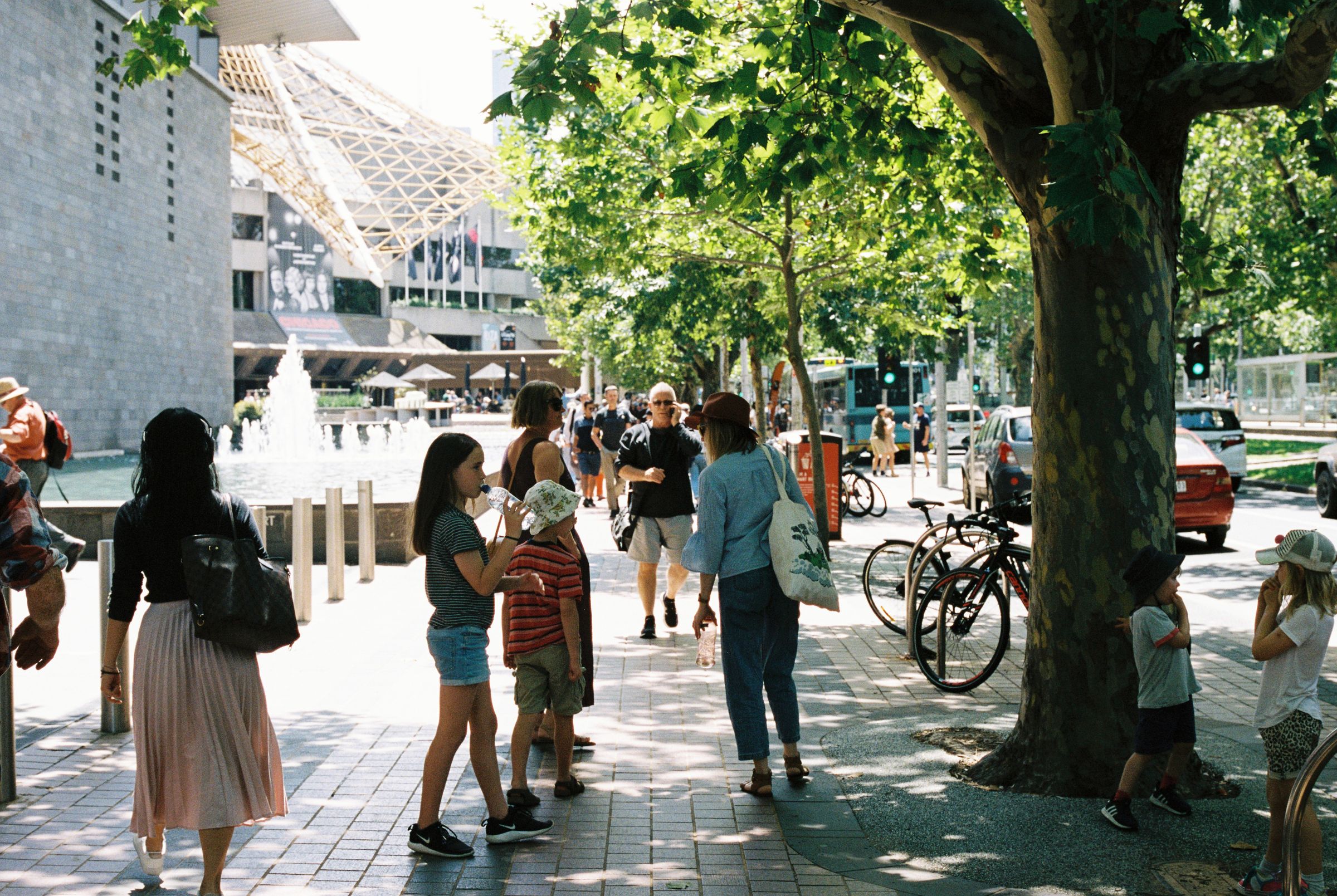 A photo of a paved street scape beside a fountain with people of all ages walking and standing. Children in hats stand in the shade of trees and bikes are parked to the side.