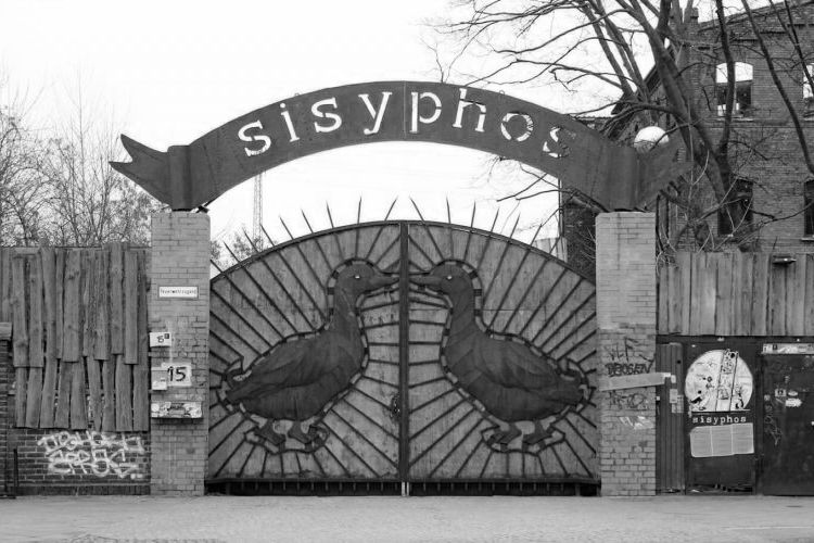 a boarded up fence with a large metal gate with ducks facing inwards on each gate and an overhead metal sign reading sisyphos and an abandoned-looking brick factory in the background