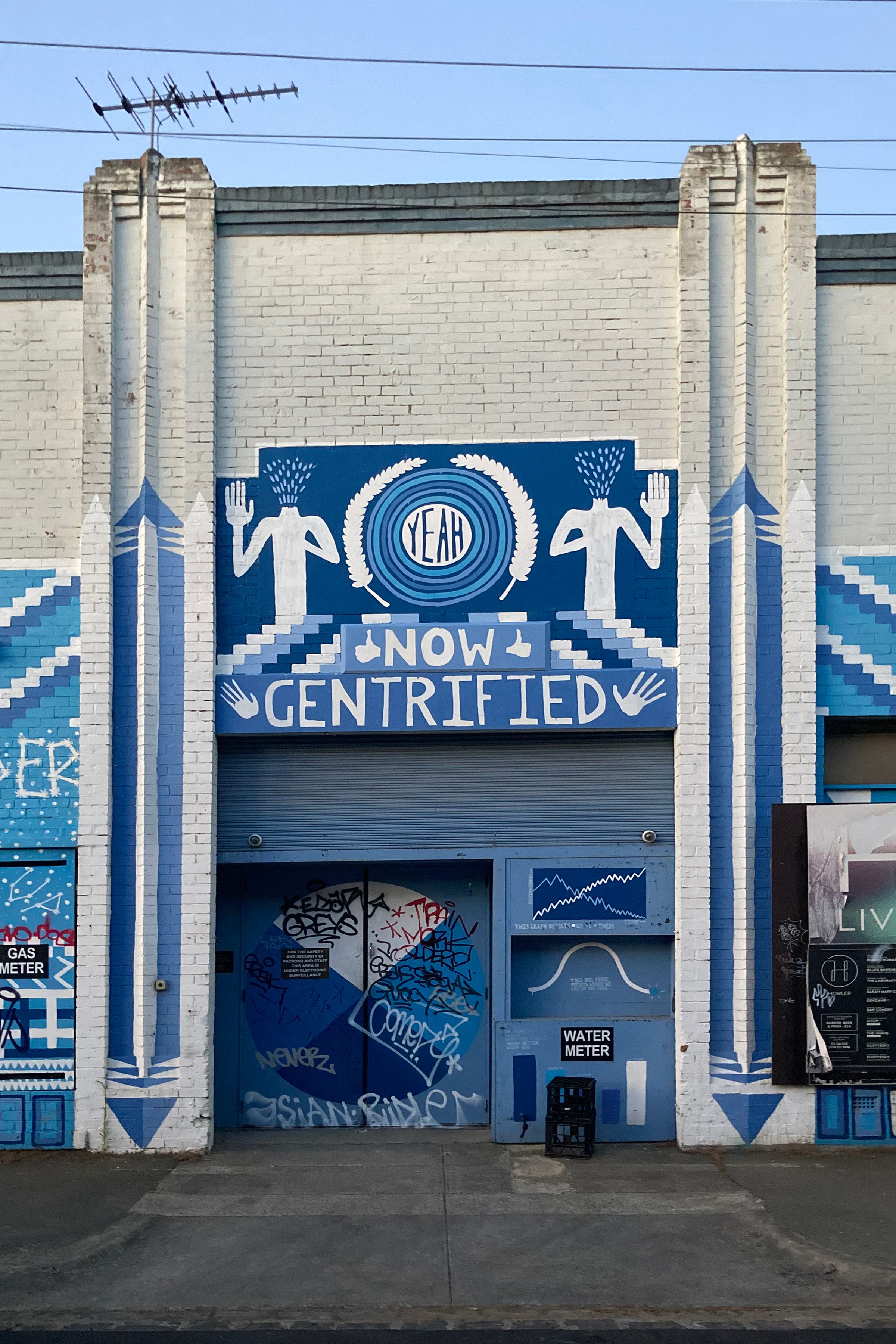 A blue and white mural painted on a warehouse building's entry with 'now gentrified' written at the top