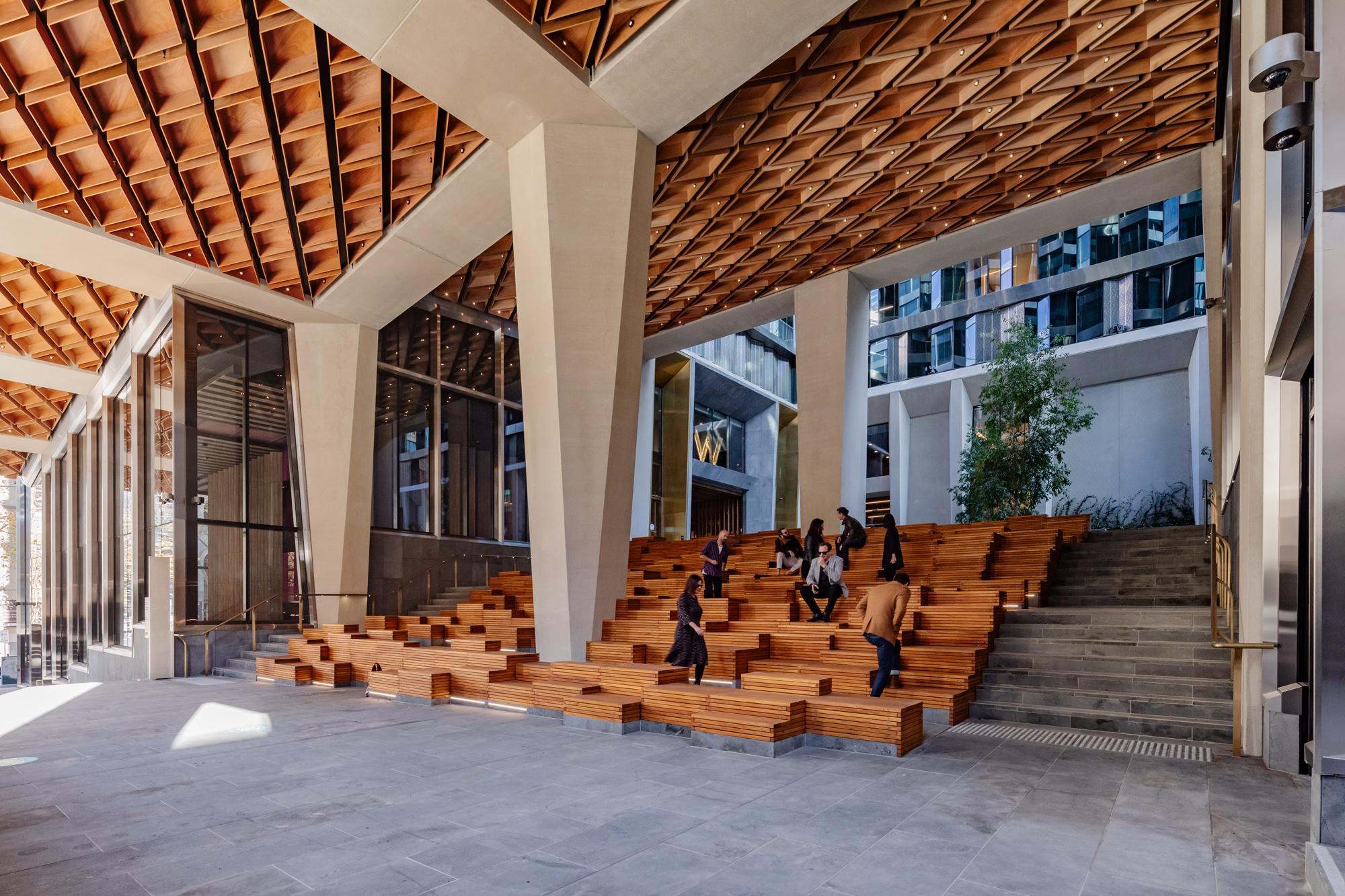 Eight people sitting or walking around wooden podium-style benches in an undercover atrium with concrete pillars and honeycomb ceiling