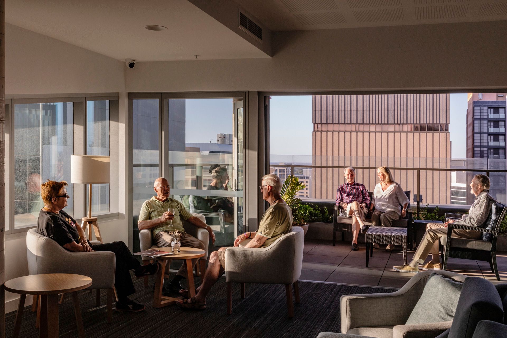 Photo of communal living space showing interior spaces connecting to a large external balcony. People are seated together and talking.