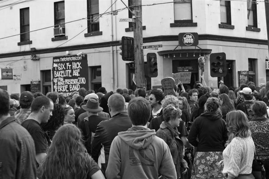 A crowd stands on the street outside a corner pub building with a person holding up a sign in the shape of a guitar that reads save live music