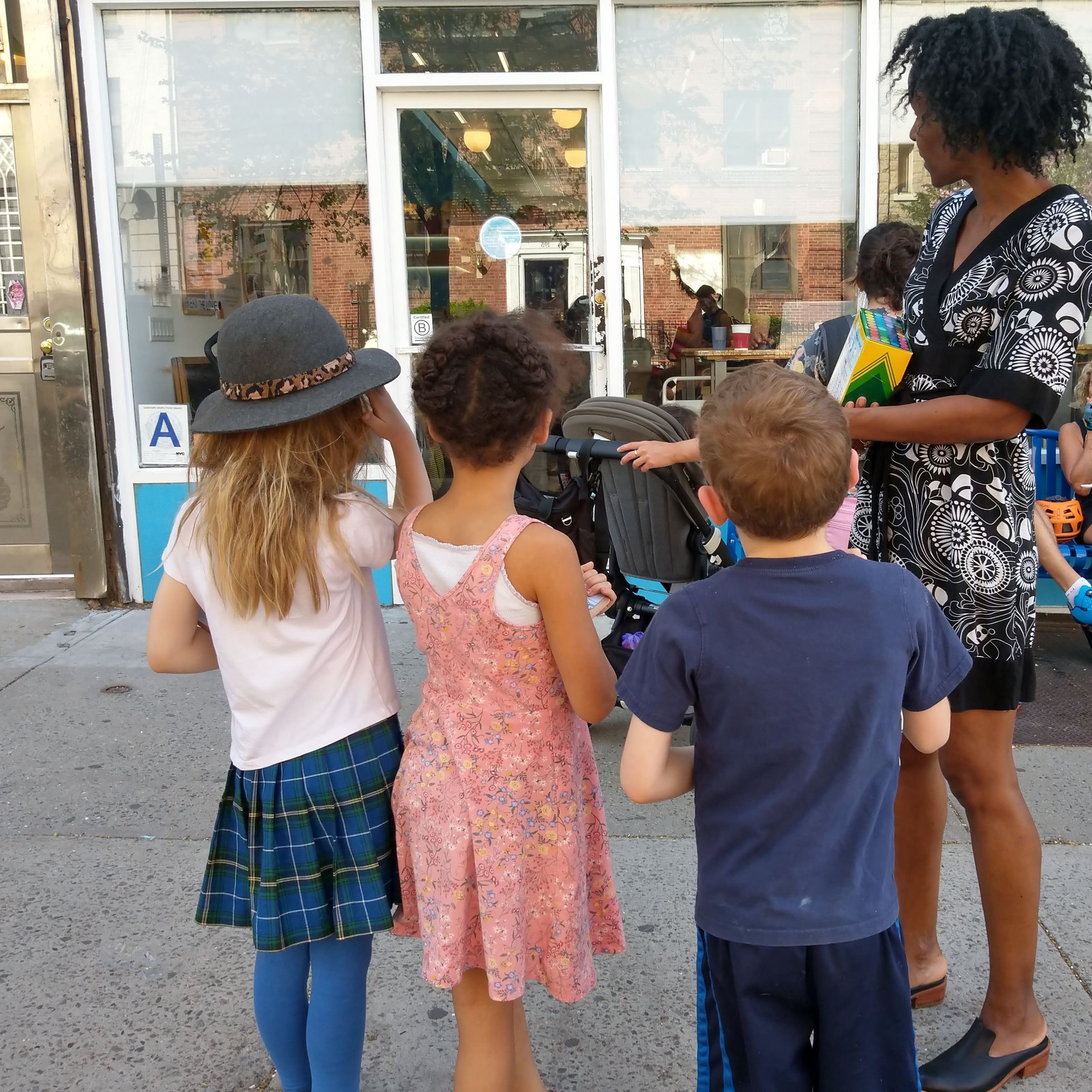 A woman stands in front of a storefront with three children who are all looking at the storefront window