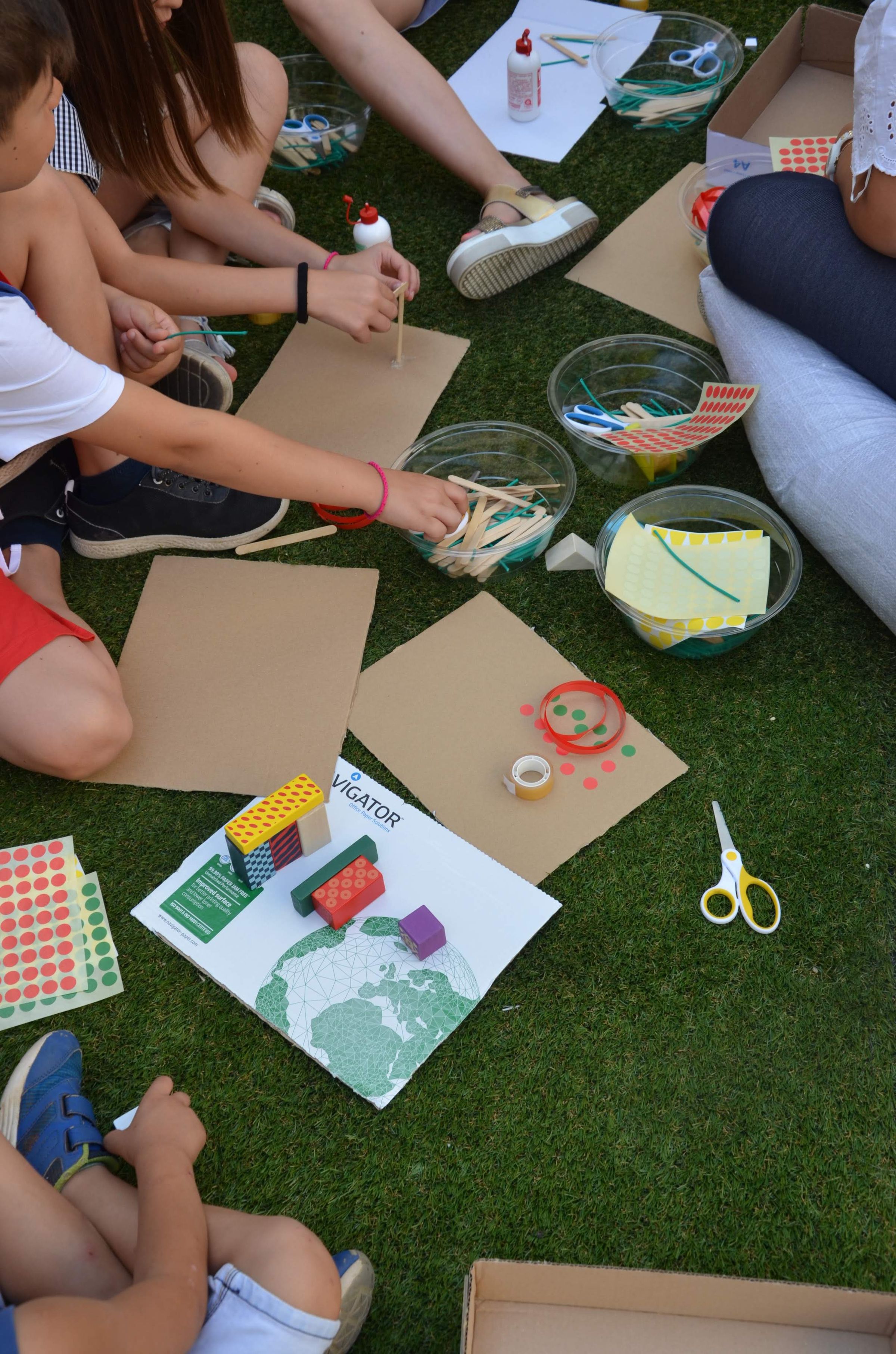 A close up of children's hands and legs, seating on synthetic grass and creating craft from coloured stickers and paddle-pop sticks on brown paper