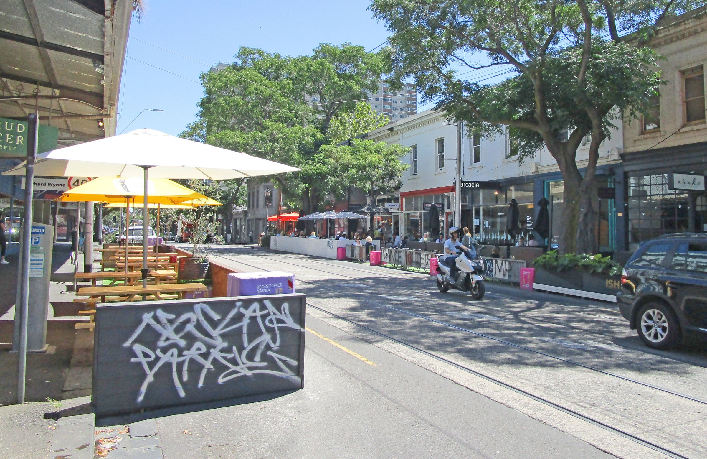 A retail street with continuous parklets on either side of the street with fencing, tables and umbrellas. A car and motorcycle drive down the tree-lined street which also has a tram line.