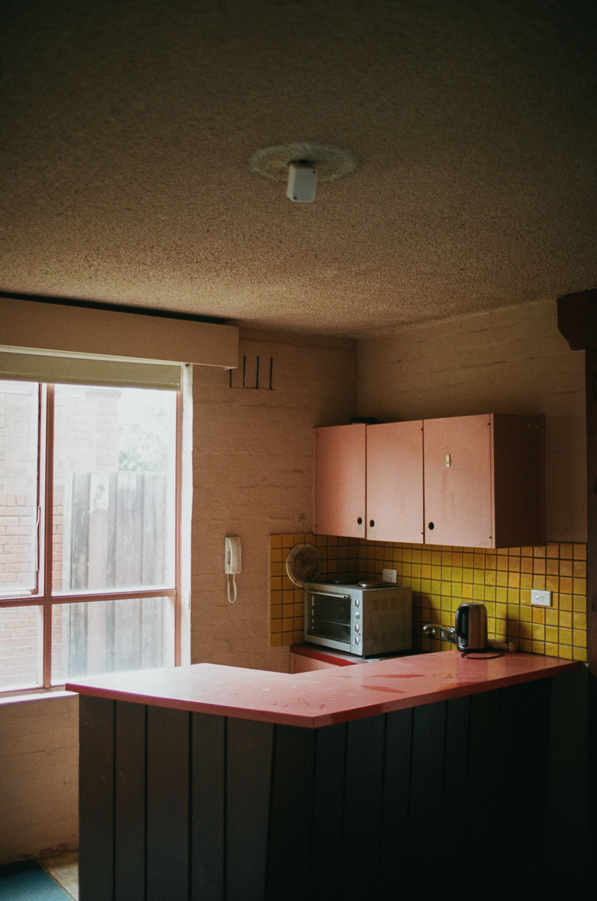 Photograph of the existing kitchen in one of the apartments, with original yellow tiles and a pink benchtop and cupboards. 