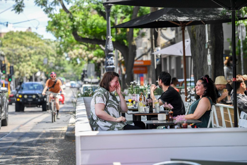 Photograph of people sitting outside in a parklet in front of Archie's all day cafe on Gertrude Street in Fitzroy. The parklet is built over car parking spaces along the street and provides a wooden barrier between the customers of the cafe and the cars and cyclists. The parklet has cafe tables where people are sitting and enjoying food and beverages.