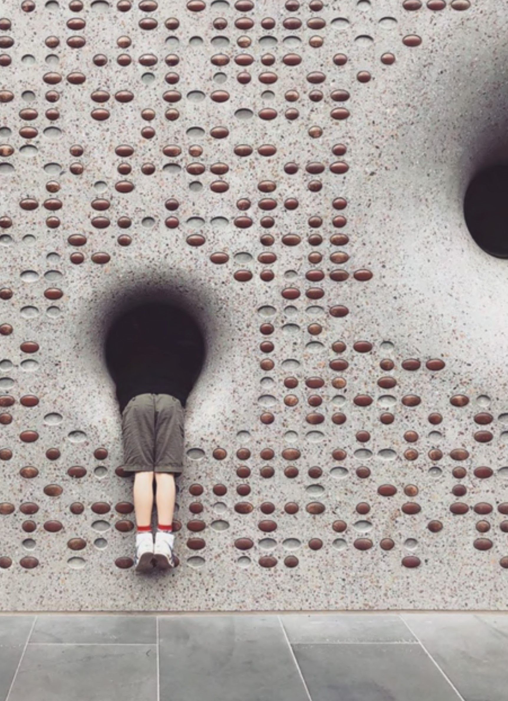 A grey concrete building wall with small tactile dots and large hollow circles with a child's playing in one circle by leaning through with its legs hanging out.