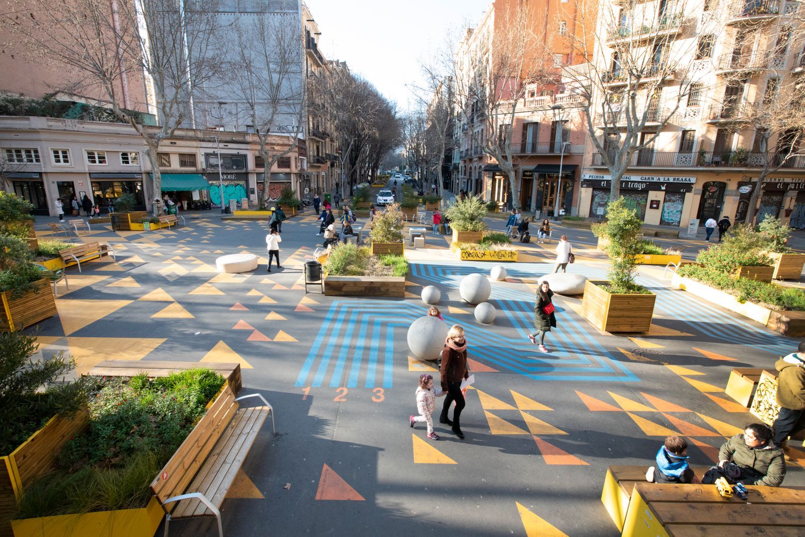Photograph of people using the pedestrian only open space provided through the superblock model. Kids are roller-blading, there are chess tables and people sitting enjoying the sun.
