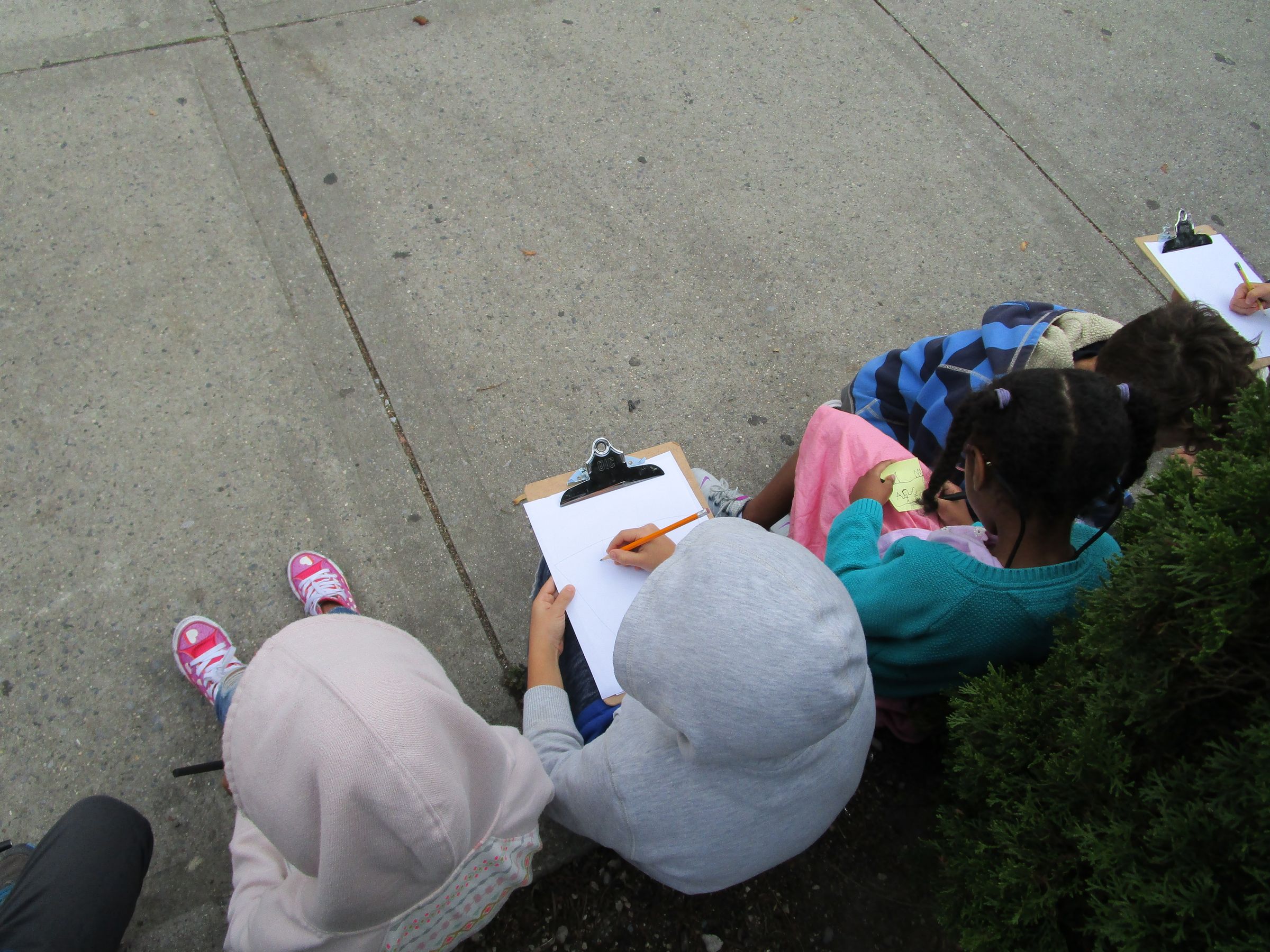 A birds-eye photograph of the top of four childrens heads who are seated on a concrete footpath writing on white paper attached to clipboards