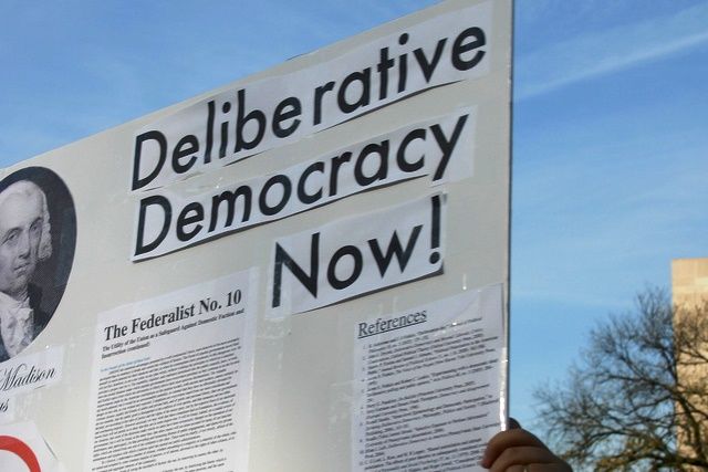 A close up pat of a hand-made placard held up to the sky with the words Deliberative Democracy Now! 