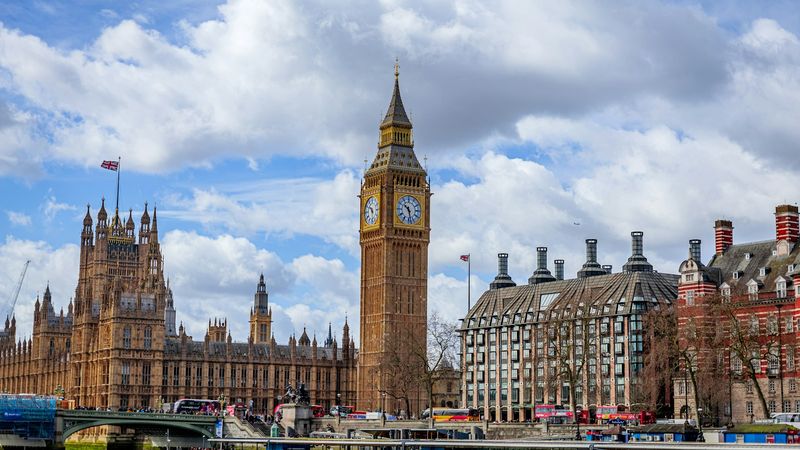 The Elizabeth Tower and Houses of Parliament in Westminster, London, viewed from across the River Thames under a partly cloudy blue sky