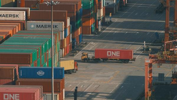 Stacks of colourful shipping containers at a busy port terminal, with a lorry carrying a red container between the rows and orange gantry cranes on the right