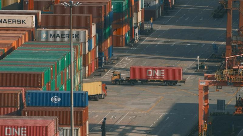 Stacks of colourful shipping containers at a busy port terminal, with a lorry carrying a red container between the rows and orange gantry cranes on the right
