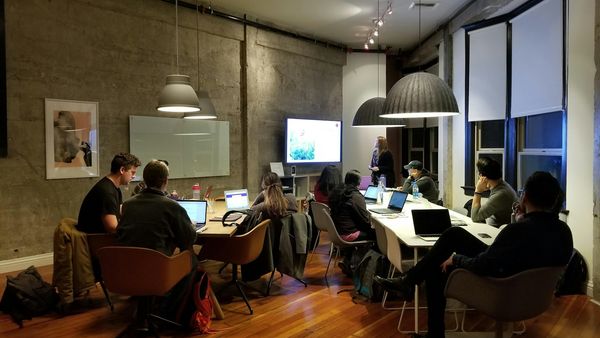A group of adults seated at tables with laptops watching a presenter speak beside a screen during a training session