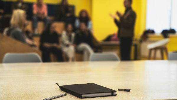 A notebook and pen on a table in the foreground, with a facilitator presenting to a seated group of professionals in a training room behind