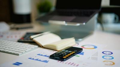 An office desk with printed charts and graphs, a smartphone calculator, a notebook, keyboard and laptop
