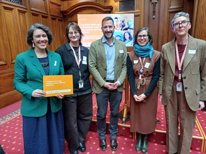 Five smiling people, one holding an orange booklet, stand in a wood-paneled room with a screen displaying text about health and wellbeing.
