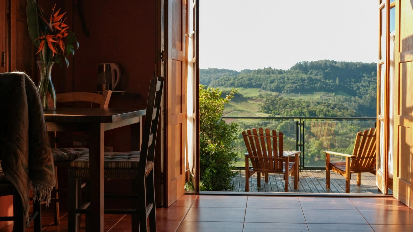 Open doors from a guest room onto a timber deck with two wooden chairs, looking over vineyards and wooded hills