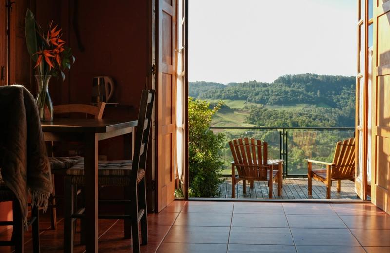 Open doors from a guest room onto a timber deck with two wooden chairs, looking over vineyards and wooded hills