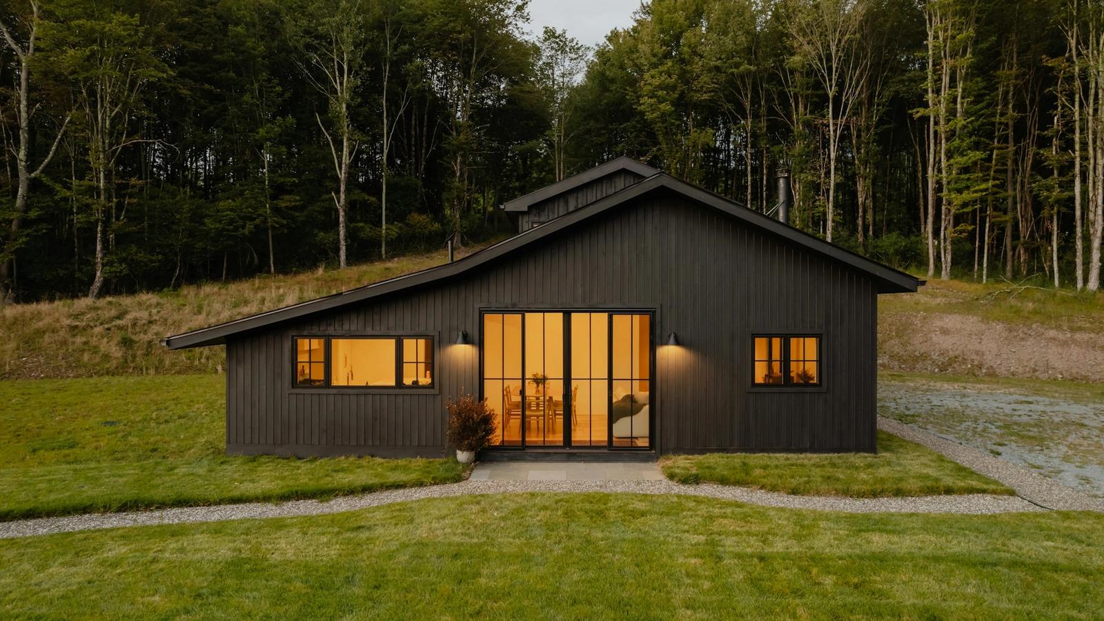 A small black timber cabin at dusk with warm light in its windows, on a lawn with tall trees behind