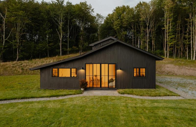 A small black timber cabin at dusk with warm light in its windows, on a lawn with tall trees behind
