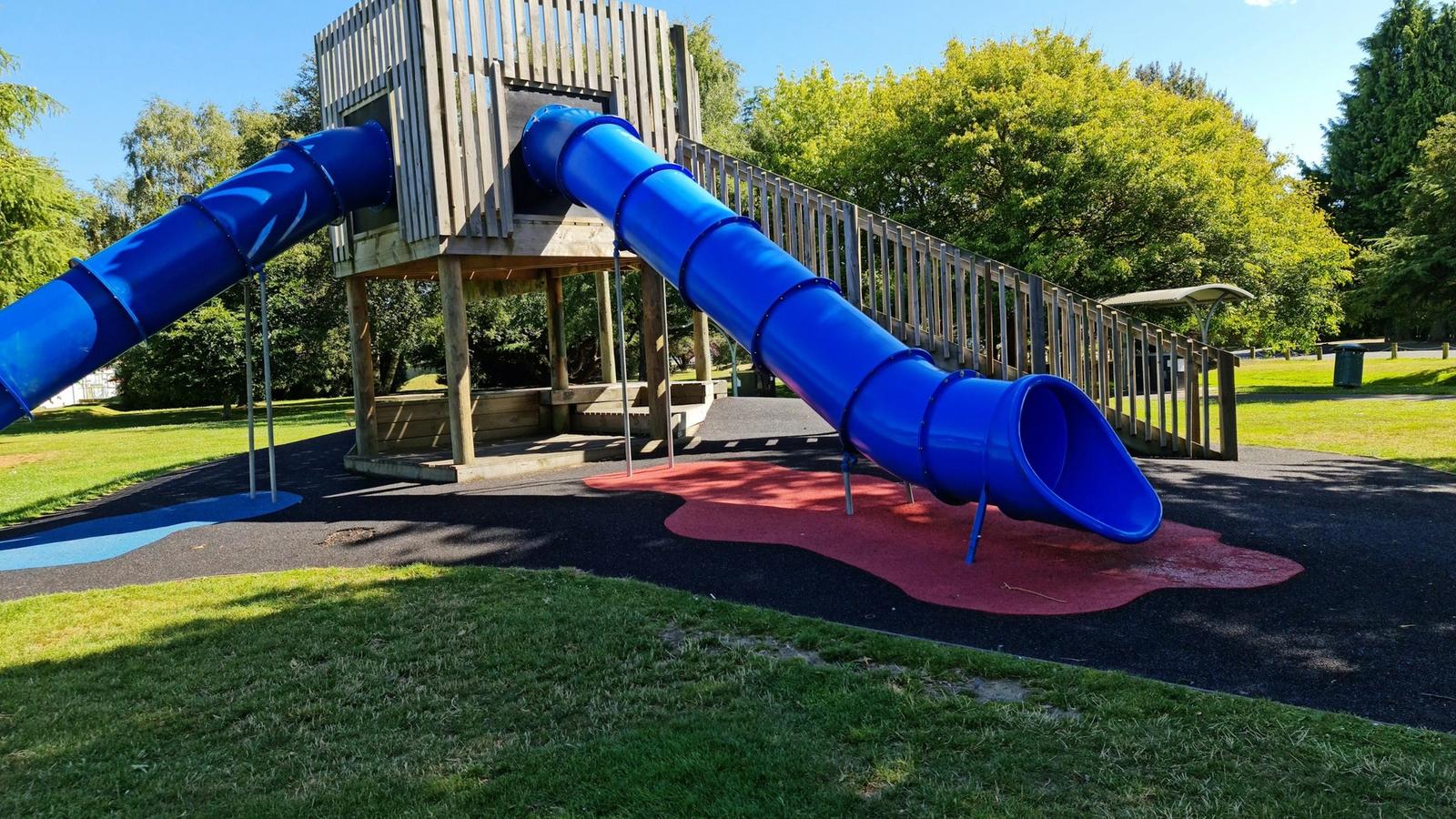 A timber play fort with two blue tube slides on a grassy lawn under a clear sky.