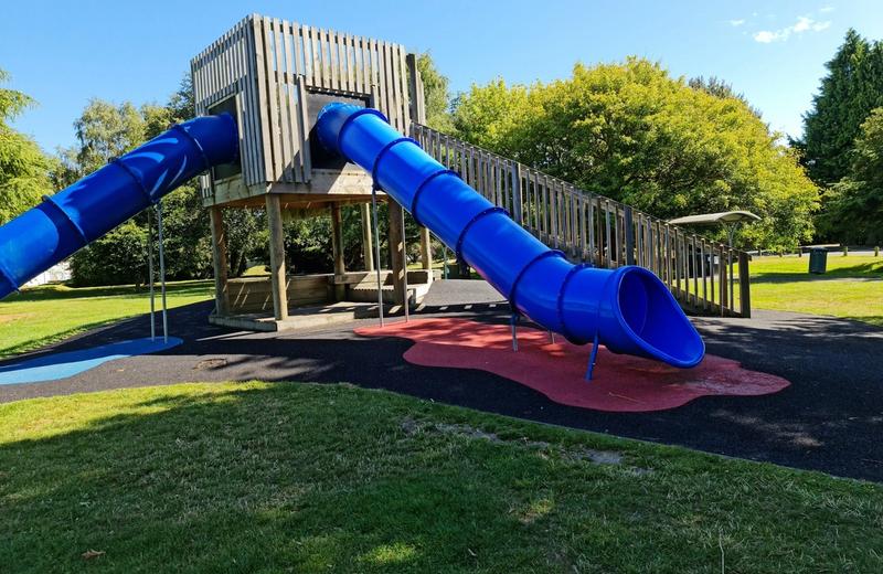 A timber play fort with two blue tube slides on a grassy lawn under a clear sky.
