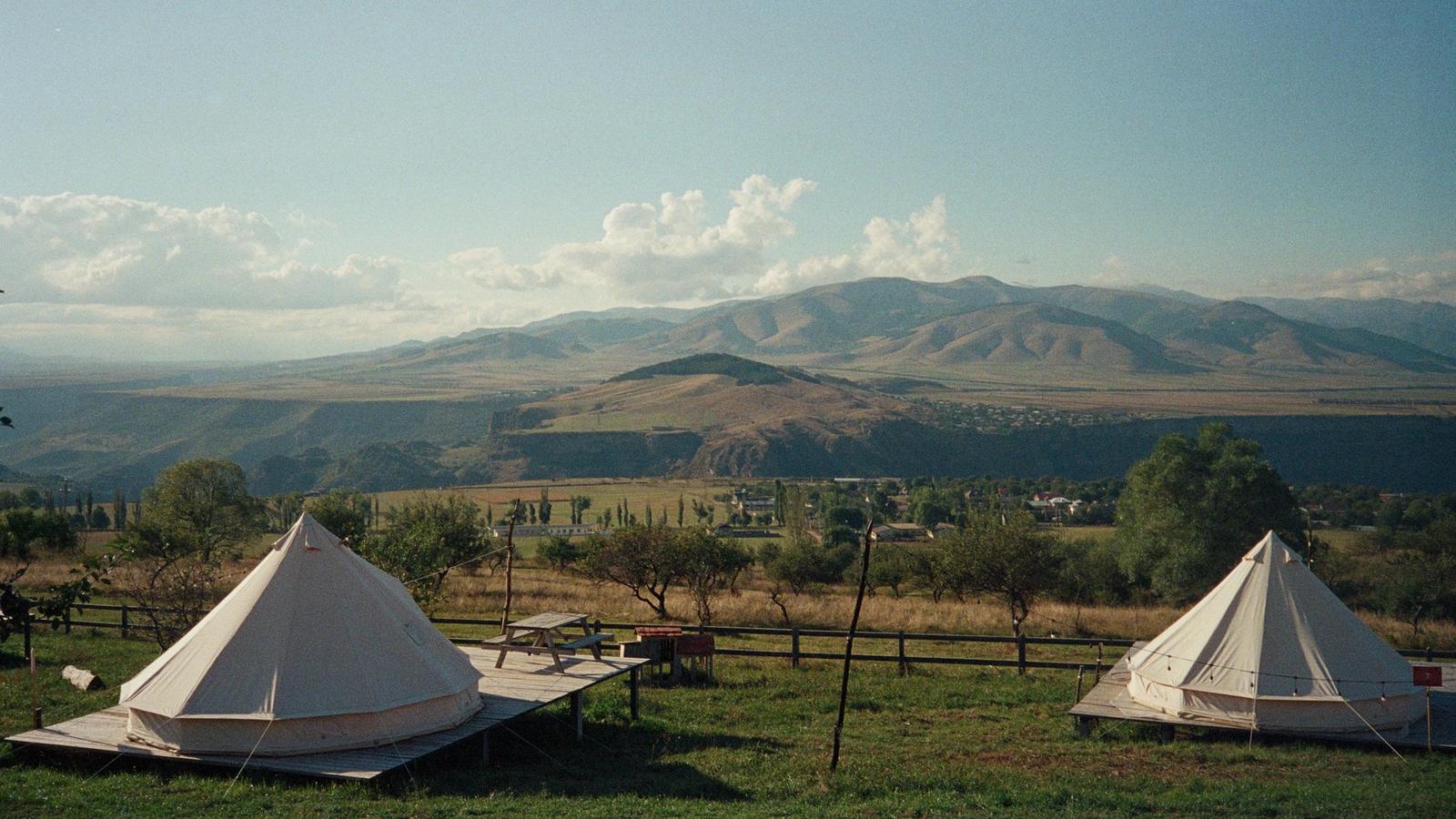 Two white canvas bell tents on timber platforms in a grassy field, with hills in the distance