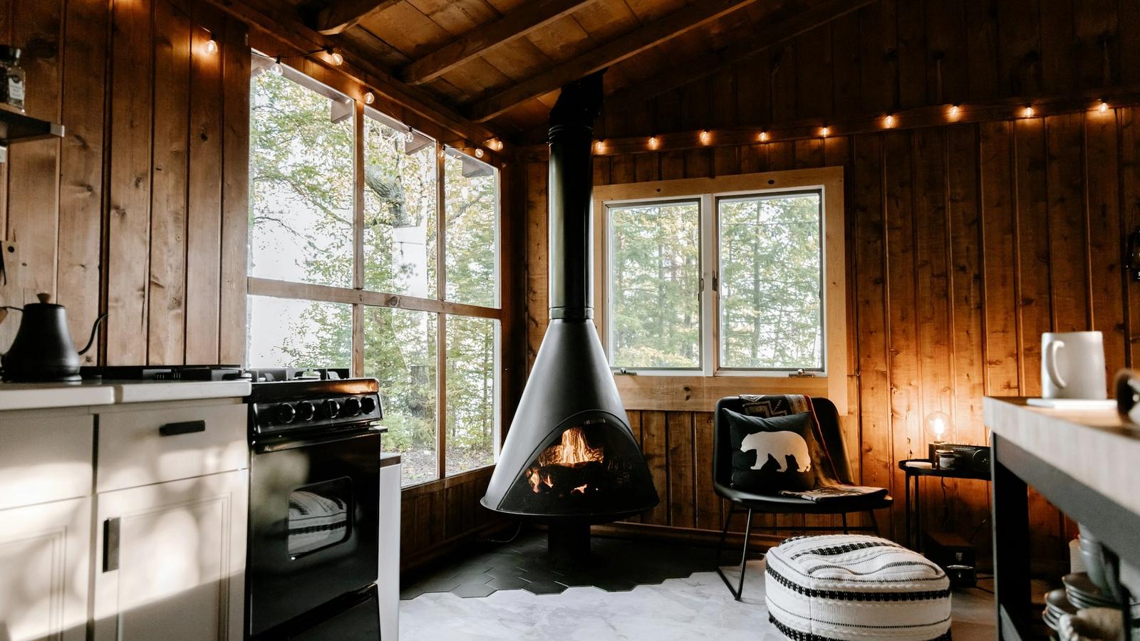 A timber cabin interior with a black hanging fireplace, big windows onto trees and warm string lights