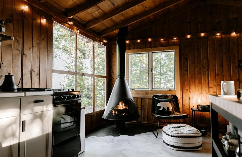 A timber cabin interior with a black hanging fireplace, big windows onto trees and warm string lights