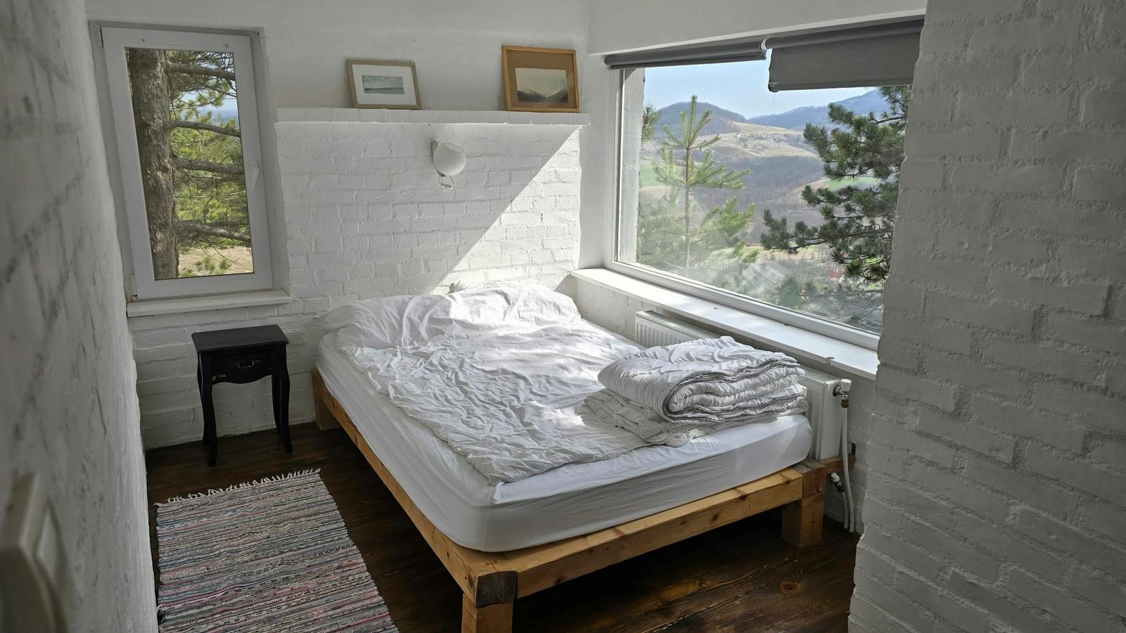 A bright white-brick bedroom with an unmade bed and a wide window looking out onto pine-covered hills