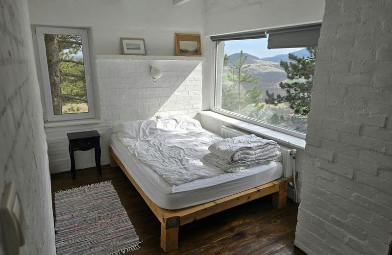 A bright white-brick bedroom with an unmade bed and a wide window looking out onto pine-covered hills