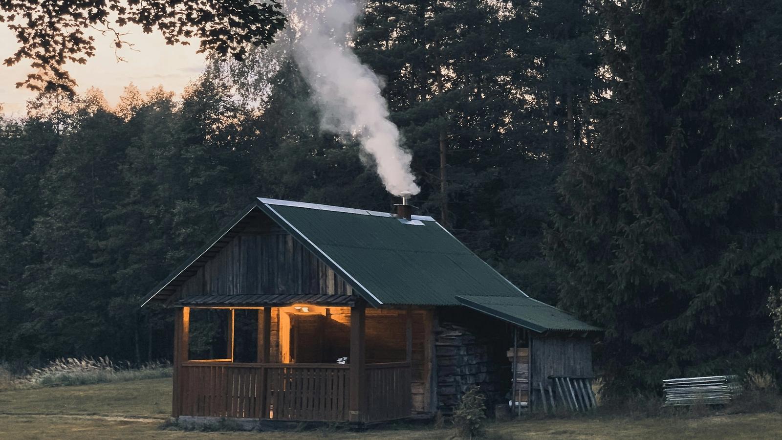 A rustic timber cabin at dusk with smoke rising from its chimney and a lit verandah, with tall trees behind