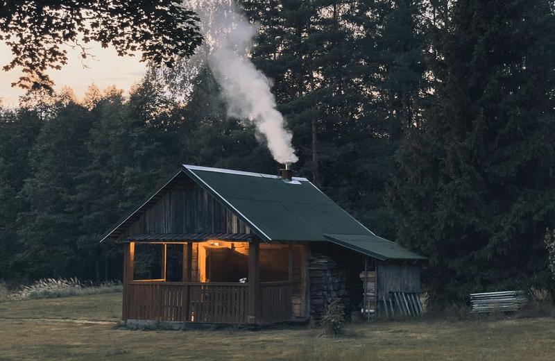A rustic timber cabin at dusk with smoke rising from its chimney and a lit verandah, with tall trees behind